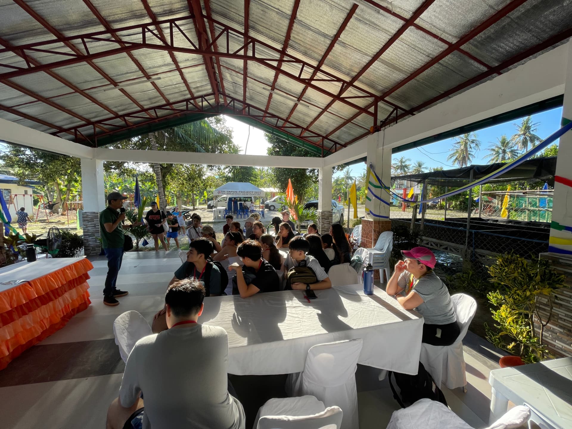 Students listening to a speaker under a covered pavilion in the village